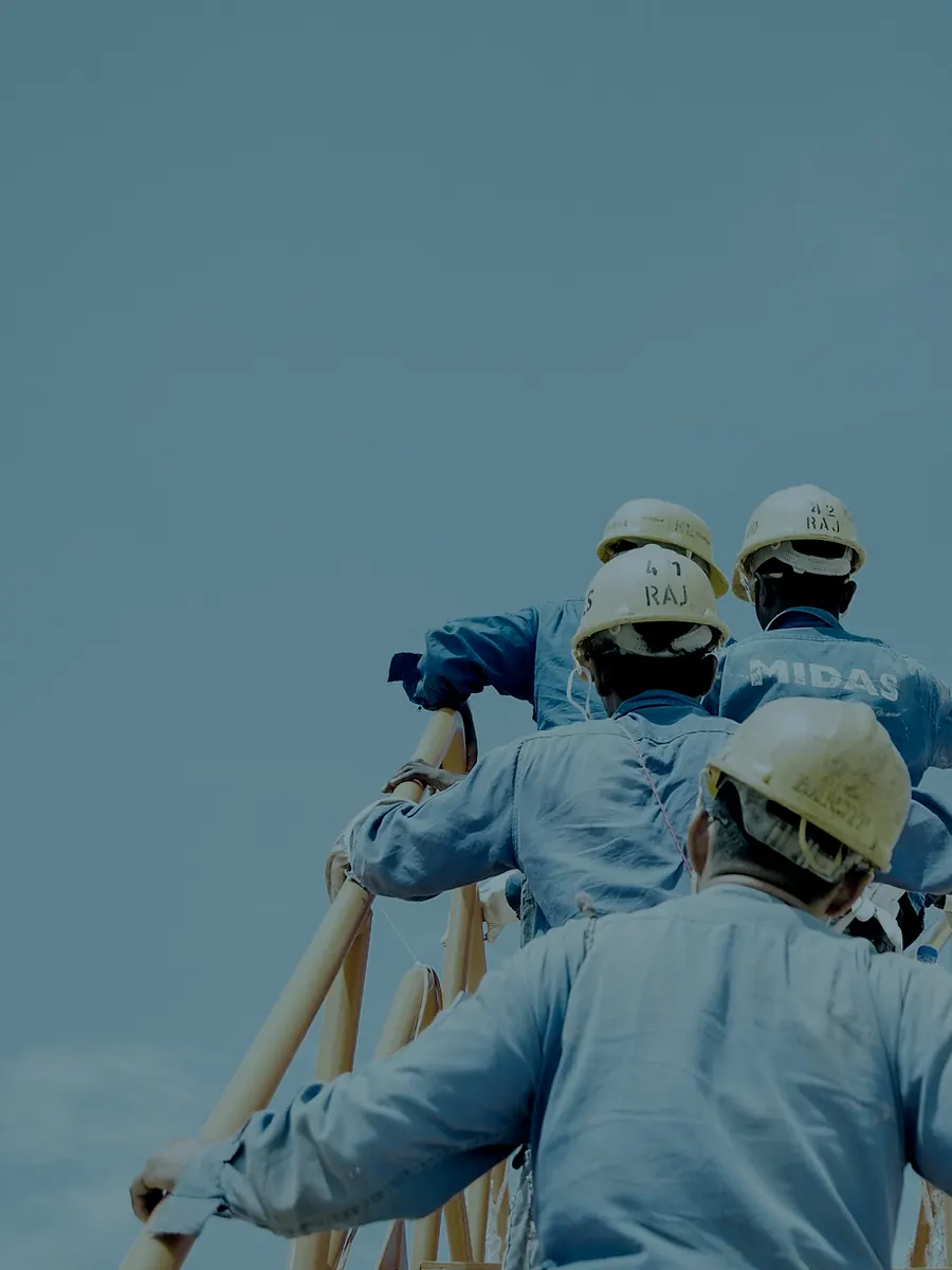 Trabalhadores de capacete e uniforme, vistos de costas, subindo uma estrutura metálica de obra contra o céu.