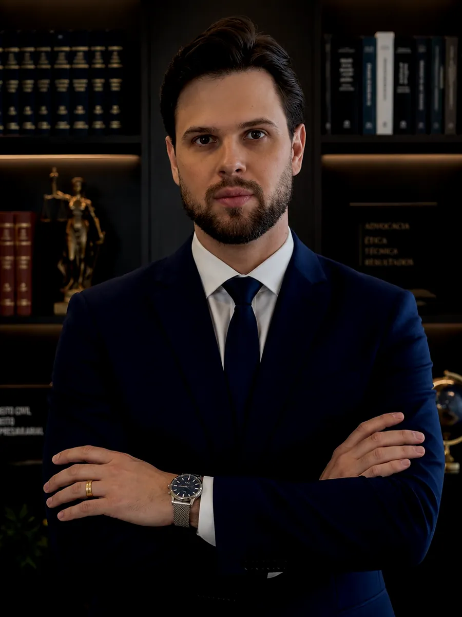 Cassio Meneghetti, advogado responsável pelo Meneghetti Advogados, de terno azul-marinho e braços cruzados, diante de uma estante de livros jurídicos.